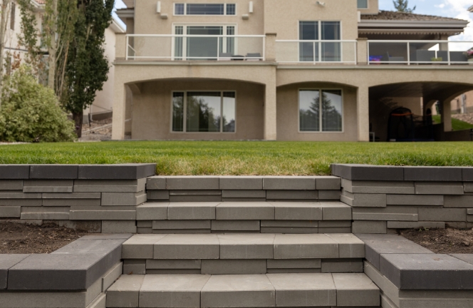 Elegant backyard with tiered stone steps leading up to a well-manicured lawn and a modern house featuring a balcony and large windows.