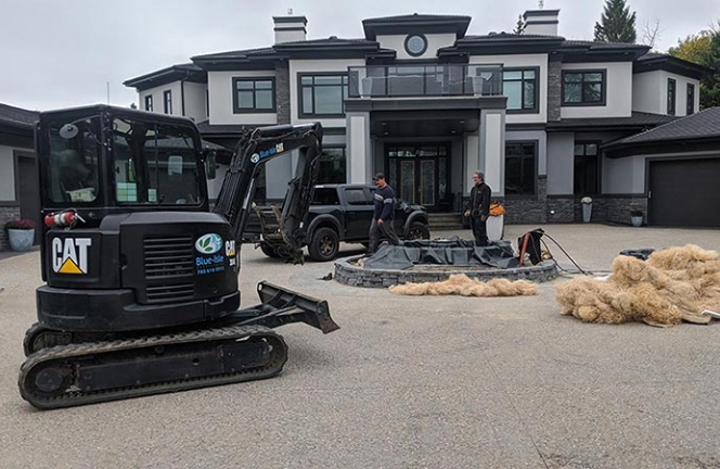 Landscaping project in progress at a modern, luxurious house featuring a large entrance with an excavator and workers installing a water feature or pond in the front yard.