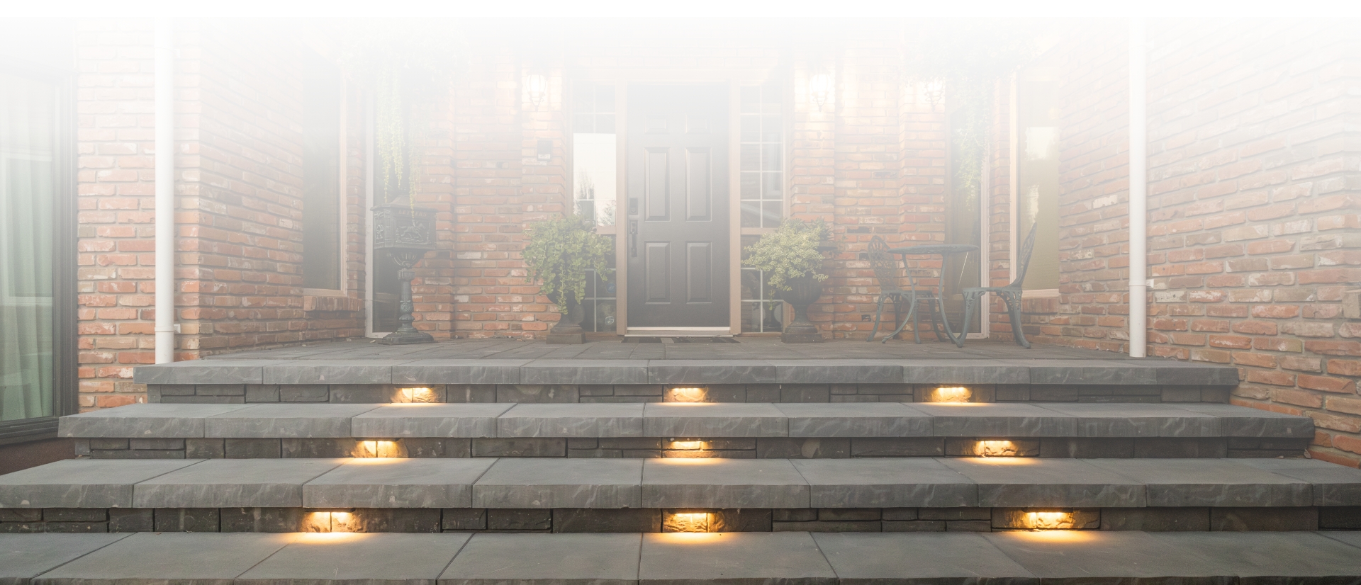 Residential home in Edmonton showcasing a hardscape design by a professional contractor, featuring illuminated stone steps leading to a dark front door, complemented by red brick walls, potted plants, and patio furniture.